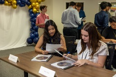 Twoyoungwomensitatatablereviewingpapersduringaclassroomorworkshopsessionwhileotherstudentsconverseinthebackgroundnearblueandgoldballoondecorations