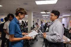 Twoyoungmenstandfacetofaceholdingpapersinaclassroomsettingsurroundedbyotherstudentsengagedindiscussionatlongtables