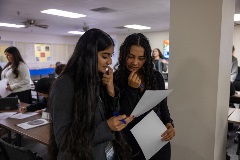 Twoyoungwomenstandclosetogetherinaclassroomreadinganddiscussingapaperwiththoughtfulexpressions