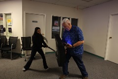 Ayoungwomanstrikesapaddedshieldheldbyanolderinstructorwearingablueshirtduringadefensivetacticsdemonstration